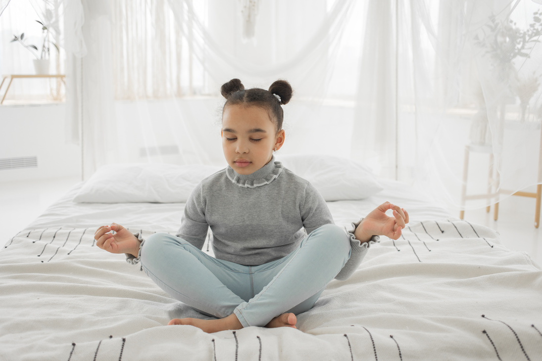 Calm black kid meditating with closed eyes and mudra hands on bed